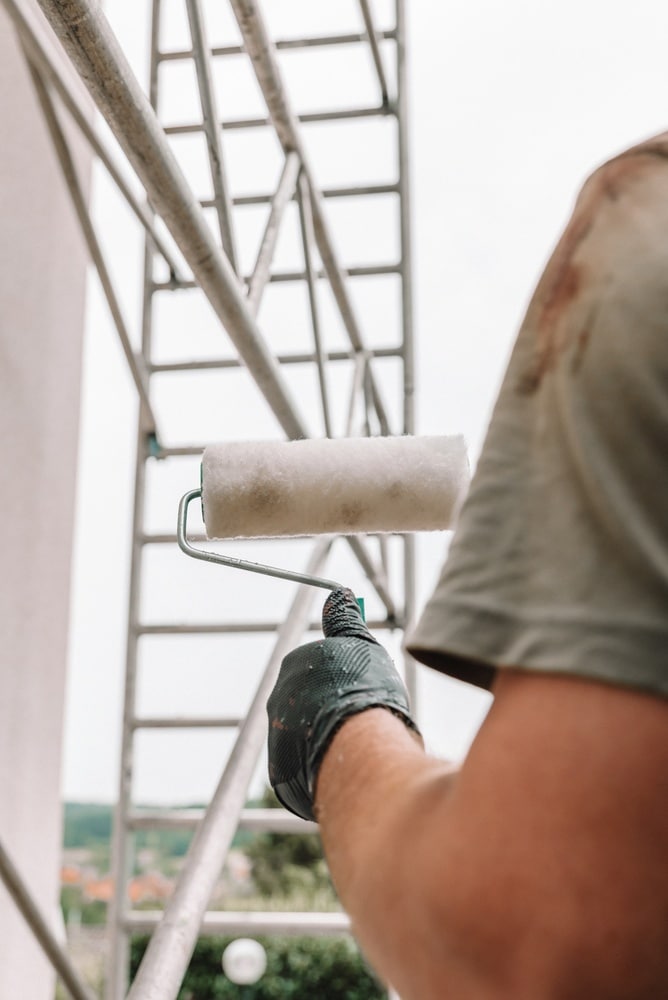 Travaux de Peinture sur Échafaudage : Guide Rénovation Peintre ganté tenant un rouleau à peinture devant un échafaudage métallique lors de travaux de rénovation extérieurs.