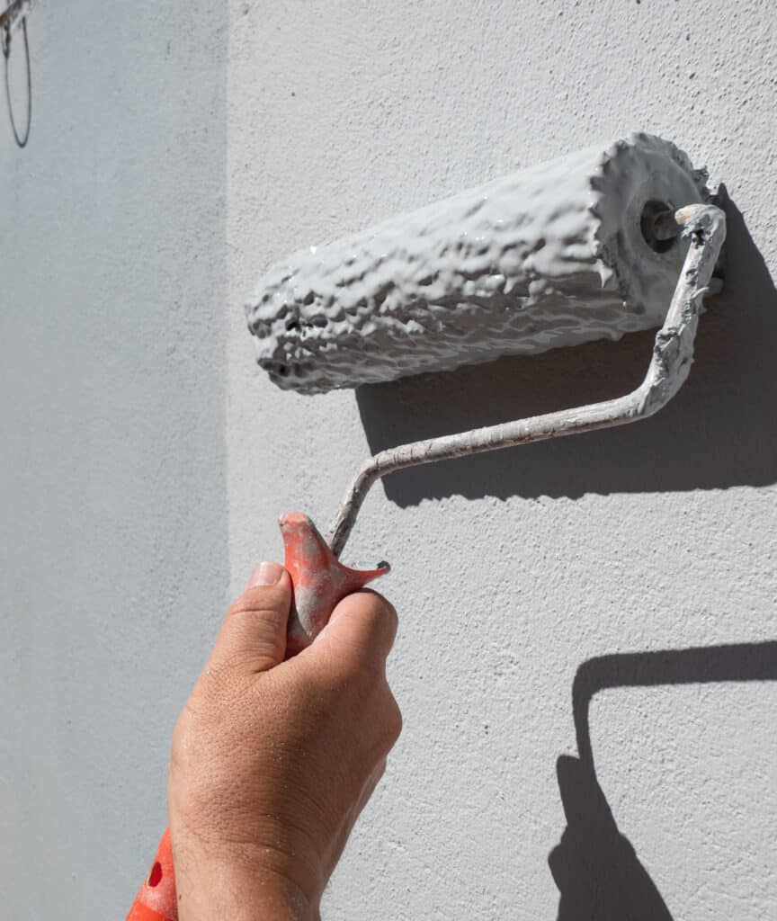 Application de peinture au rouleau sur mur extérieur Main tenant un rouleau à peinture gris chargé sur un mur extérieur crépi sous une lumière vive.