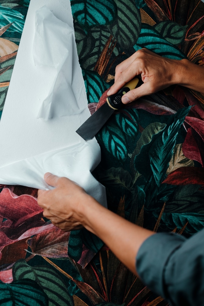Installation de papier peint aux motifs tropicaux Mains utilisant un grattoir pour enlever un film blanc d'un papier peint aux motifs de feuilles tropicales.