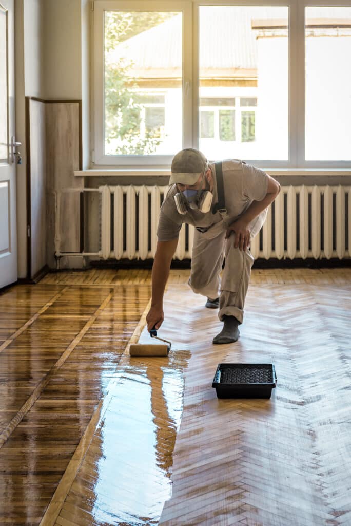 Vernissage de parquet : rénover et protéger son sol en bois Artisan appliquant du vernis protecteur sur un parquet en bois avec un rouleau dans une pièce ensoleillée.