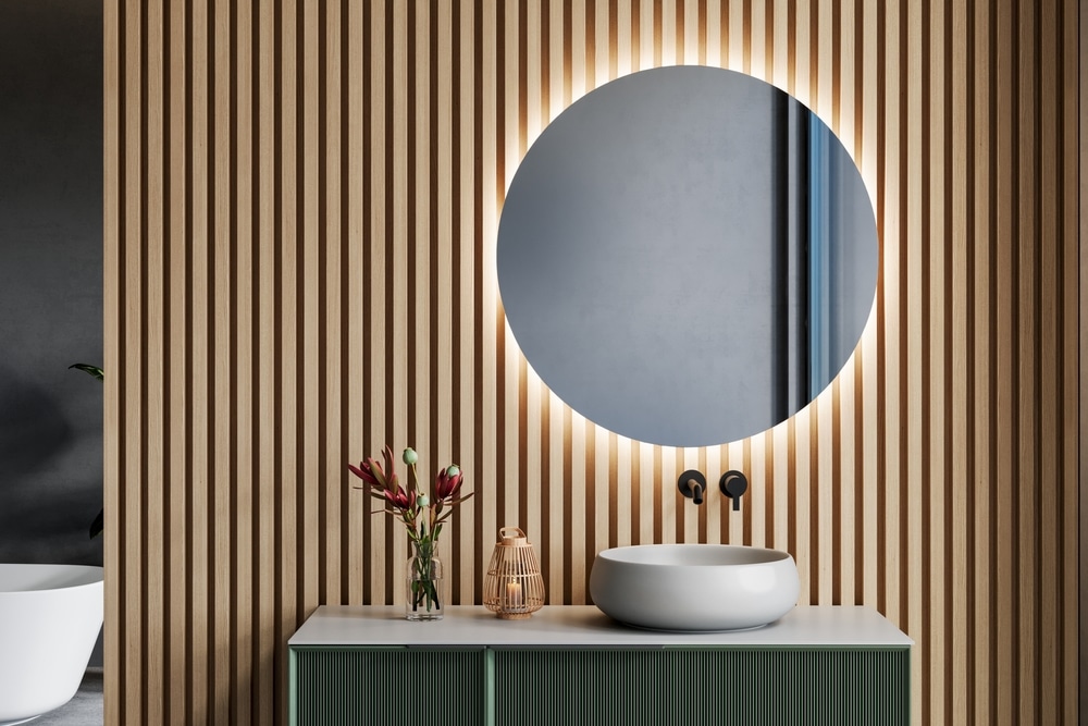 Miroir rétroéclairé et mur bois salle de bain moderne Salle de bain moderne avec miroir rond rétroéclairé, mur en tasseaux de bois, vasque blanche et meuble vert.